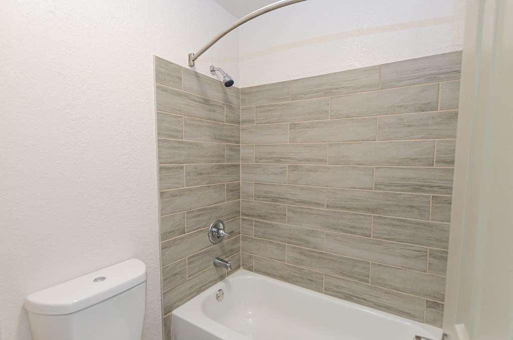 A white toilet sits next to a white bathtub in a bathroom with grey tiled walls.