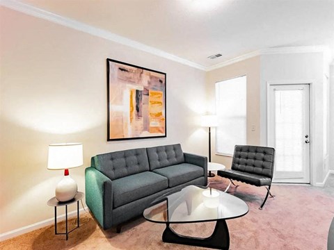 A living room with a grey couch, a black chair, a glass table and a painting on the wall.