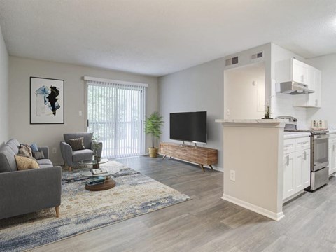 A living room with a grey couch, a wooden coffee table, a flat screen TV, and a kitchen area with white cabinets.