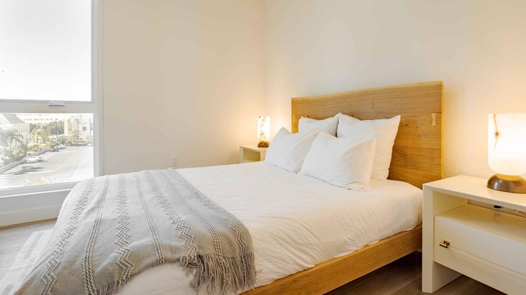 A bed with a white blanket and pillows is in the foreground with a nightstand to the right.