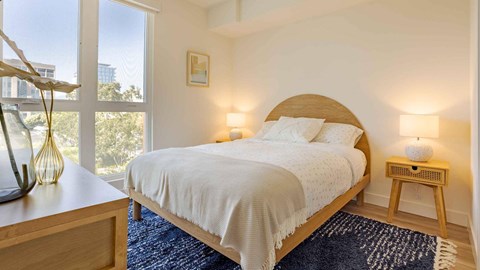 A bedroom with a bed, nightstand, and a window overlooking a cityscape.