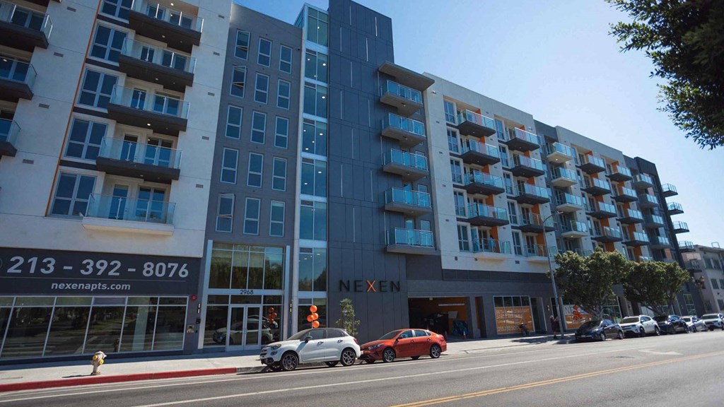 A red car is driving past a building with the name Nexen on it.
