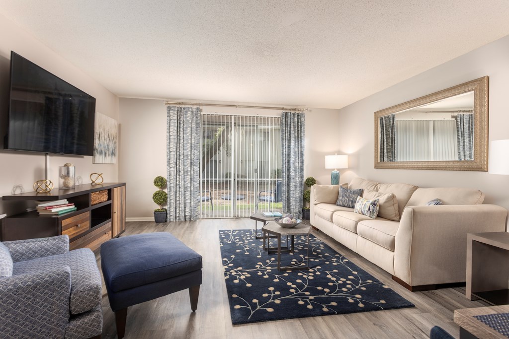 A living room with a grey chair, a blue ottoman, a white couch, a wooden coffee table, a flat screen TV, and a large mirror.