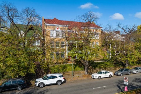 A red-roofed building is surrounded by trees and cars are parked on the street.