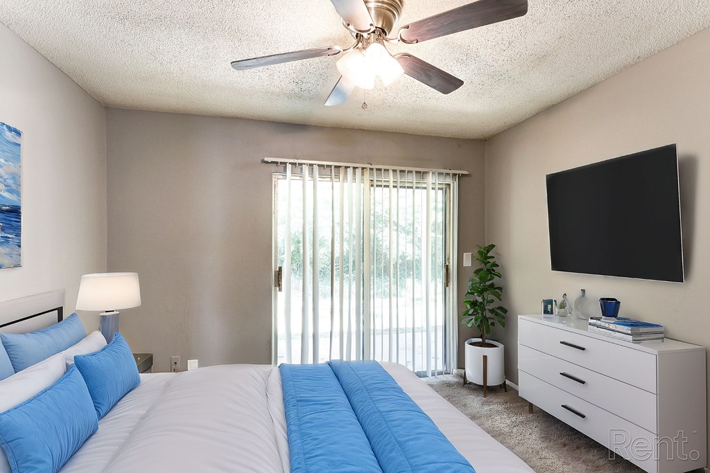 A bedroom with a bed, ceiling fan, a flat-screen TV mounted on the wall, and a sliding door leading outside.