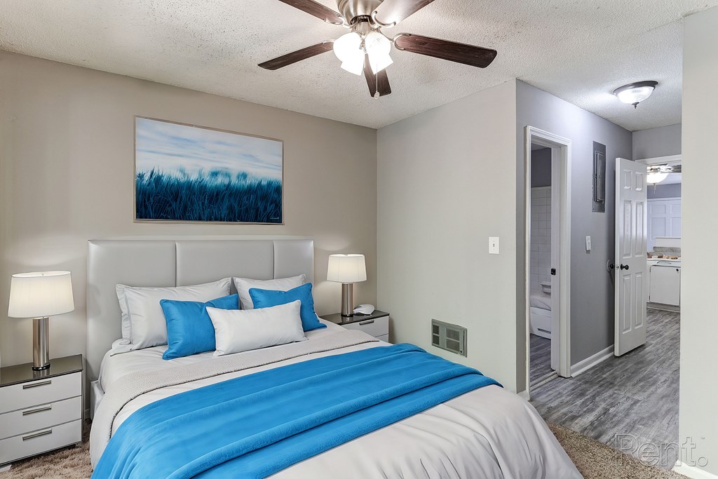 A bedroom with a bed, two lamps, a ceiling fan and a painting on the wall.