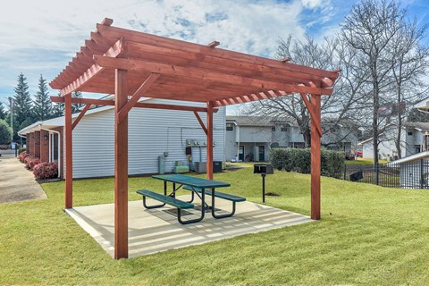 A wooden pergola with a green table is in the middle of a grassy area.