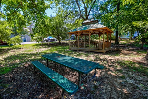 A picnic table and bench are set up in a park with a gazebo in the background.
