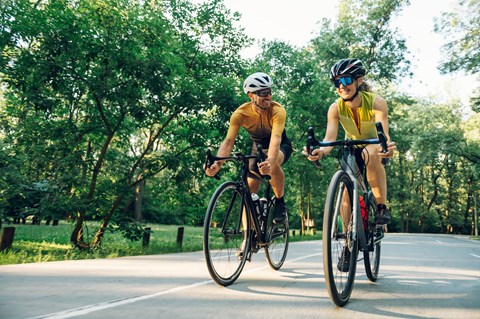 Two cyclists wearing helmets and sunglasses ride their bikes on a road.