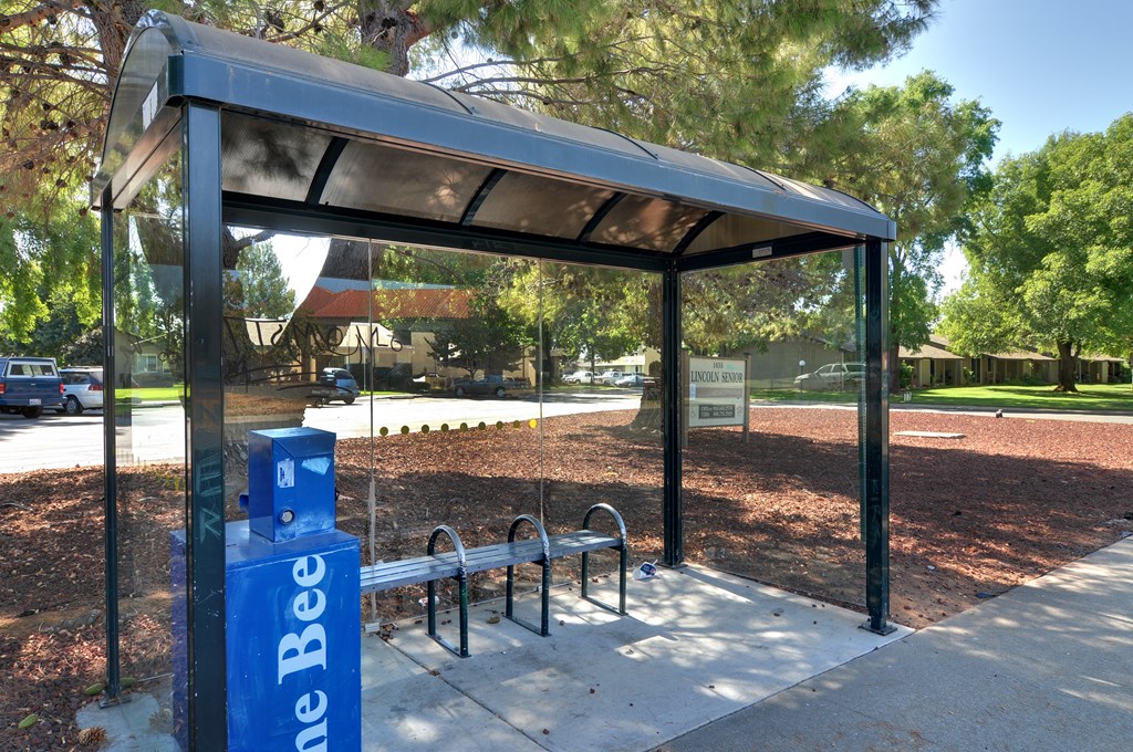 A blue and white sign that says "Bee" is on a pole next to a bench under a shelter.
