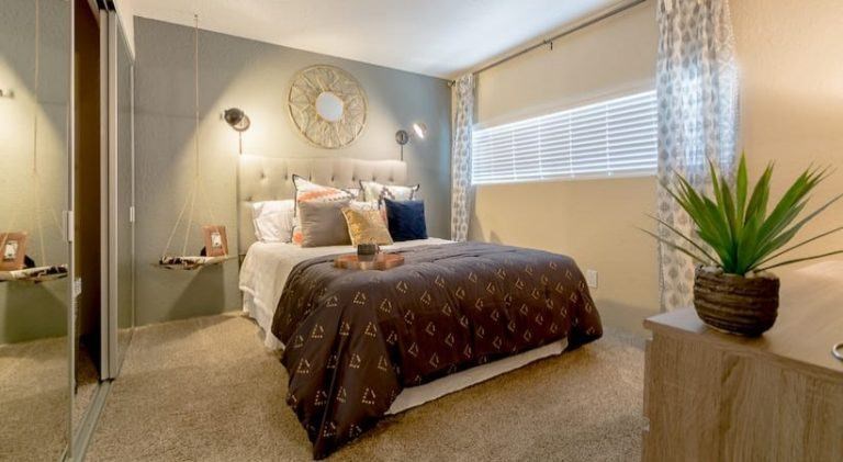 A bedroom with a bed covered in a patterned bedspread and a potted plant on the side table at Infinity on the Landing Apartments, Euless, TX