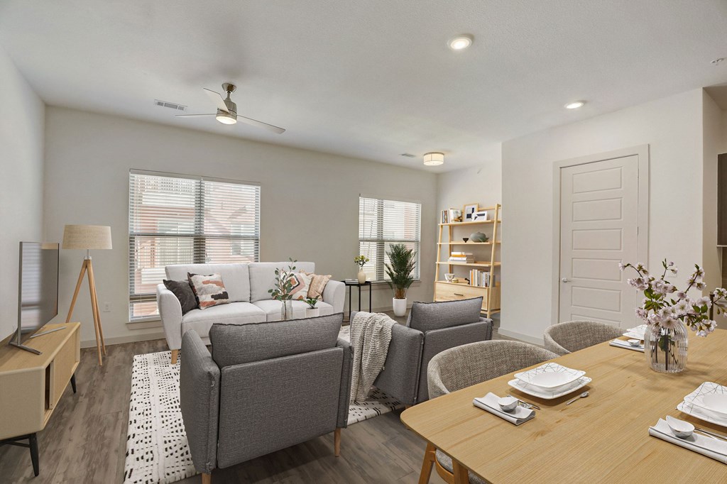 A well-lit living room with a grey couch, a wooden dining table set for two, and a bookshelf filled with books at Infinity on the Point Apartments, Dallas, Texas
