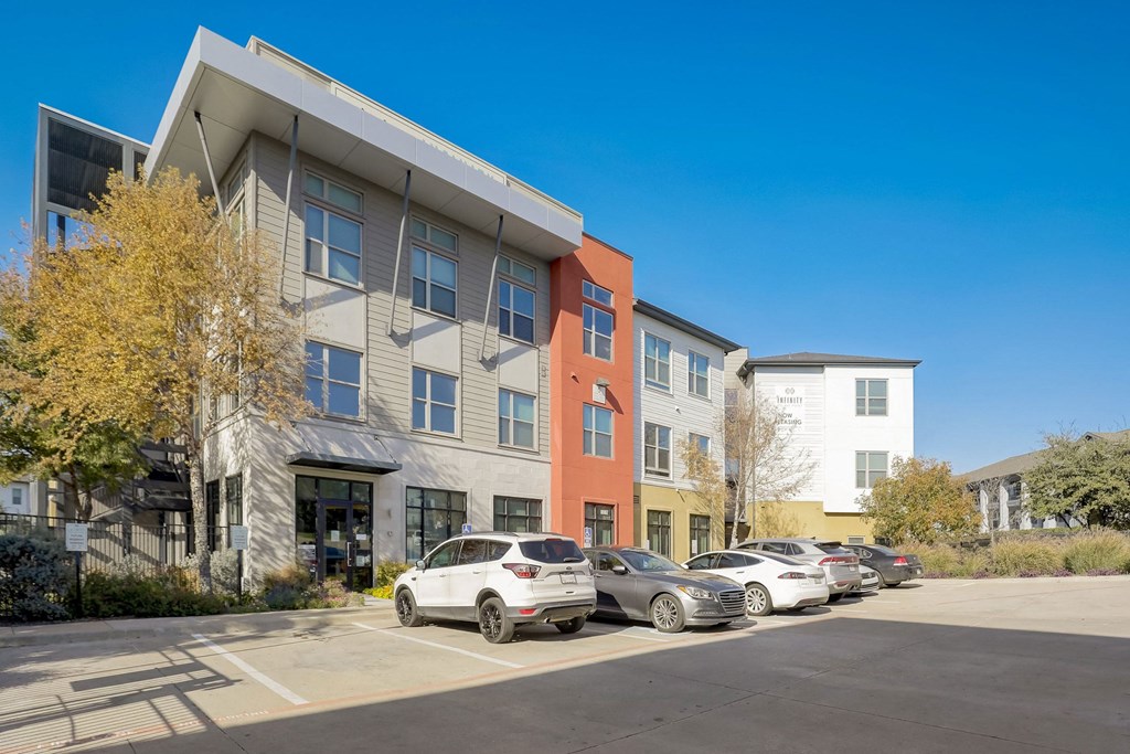 A parking lot with cars and a building with a white car parked in front at Infinity on the Point Apartments, Dallas, TX