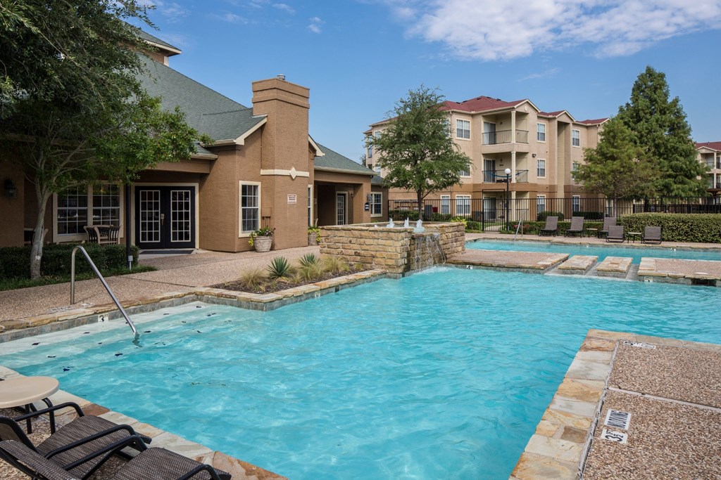 A large outdoor swimming pool with a stone border and a small patio area with chairs and a table.