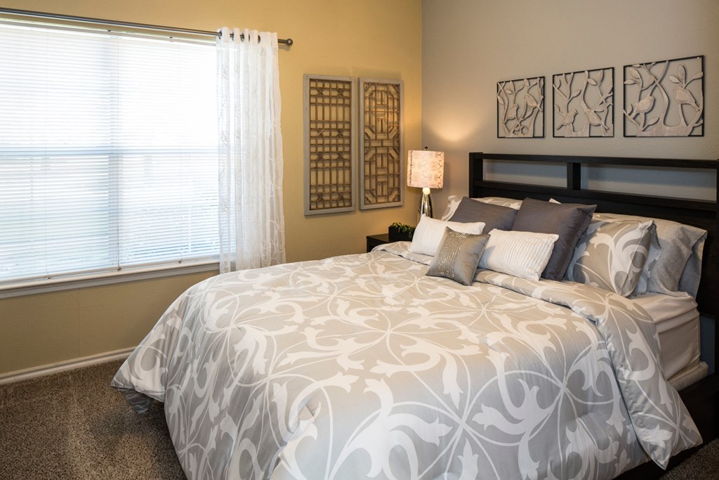 A bed with a white and grey patterned comforter sits in a room with a window and wall art.
