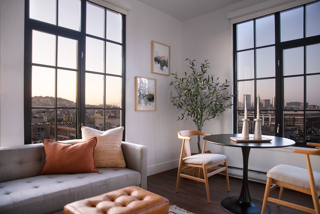 A living room with a grey couch, a round table, and a view of the city through the windows.