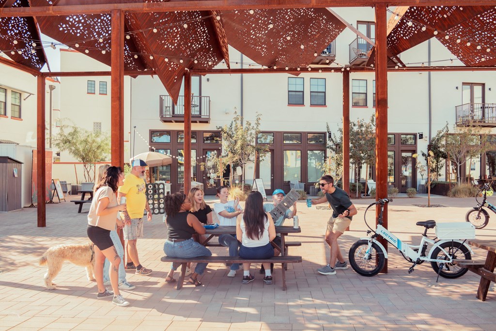 A group of people are sitting and standing under a canopy in a sunny outdoor area.