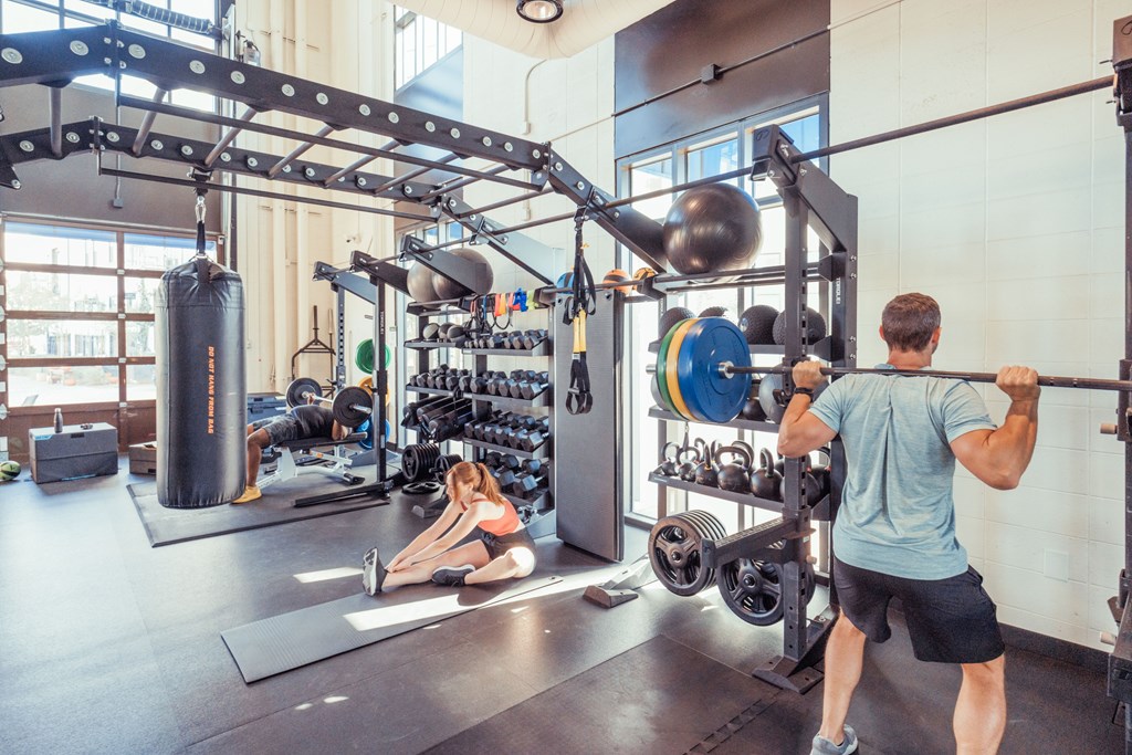 A man is lifting a barbell in a gym with a woman stretching on the floor.