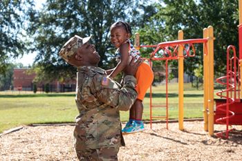 A soldier in camouflage uniform holding a child at a playground.