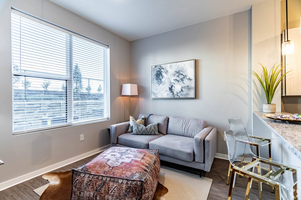 A living room with a grey couch, a patterned ottoman, and a painting on the wall.