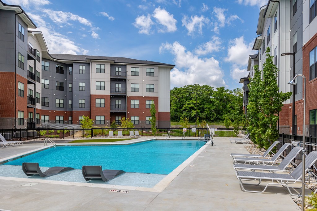 A swimming pool surrounded by lounge chairs in front of a multi-story apartment building.