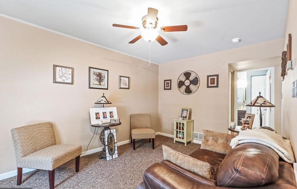 A living room with a brown leather couch, a fan, and a table with a picture frame on it.