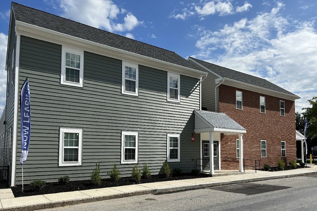 A two-story building with a grey exterior and a red brick building in the background.