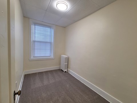 A room with a carpeted floor, a window with blinds, and a white radiator.