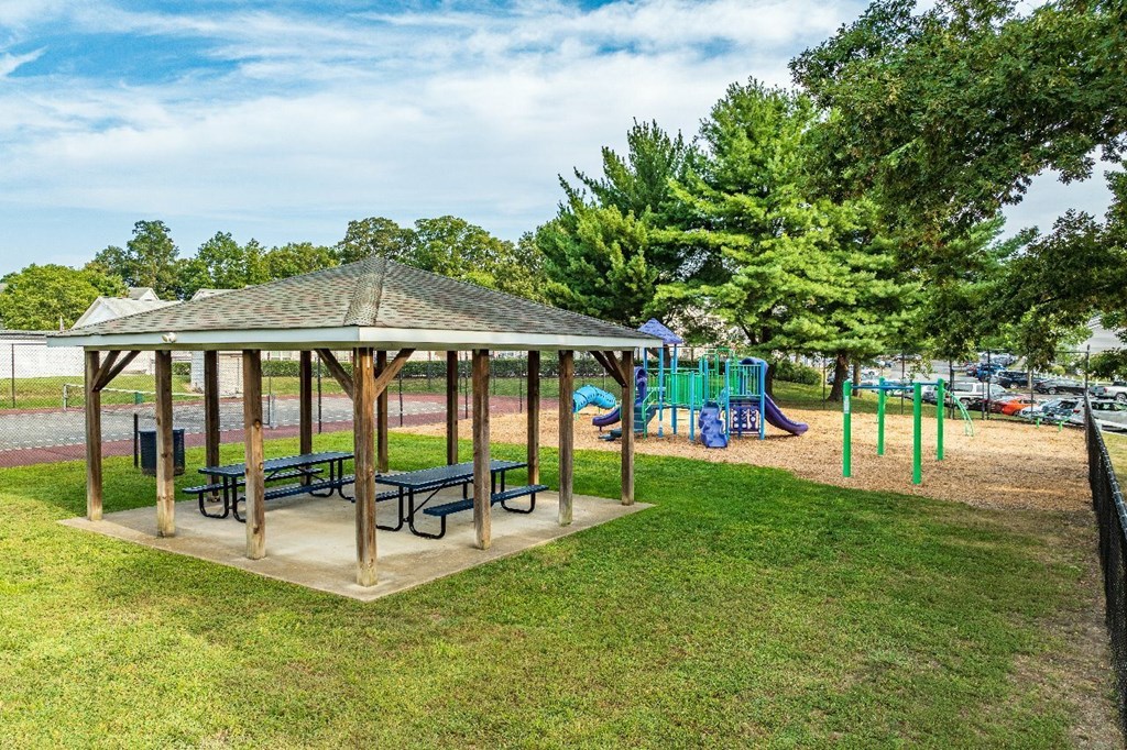 A gazebo with picnic tables is surrounded by trees and a playground.