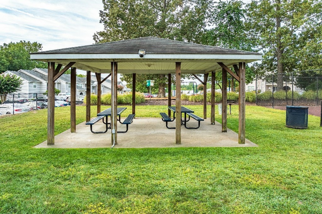 A wooden pavilion with a roof and four benches sits in a grassy area.