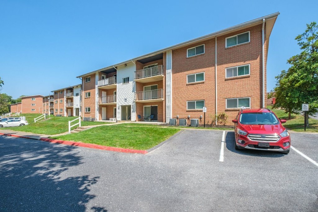A red car is parked in a parking lot in front of a brick apartment building.