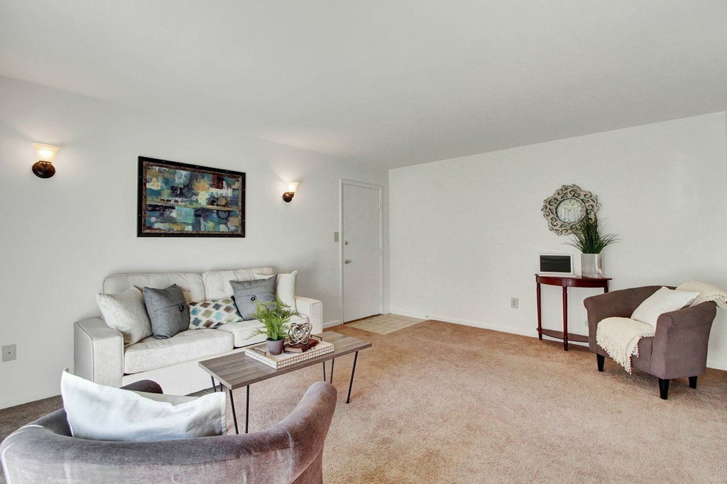 A living room with a grey armchair, a white sofa, a small table with a plant and a clock on it, and a painting on the wall.