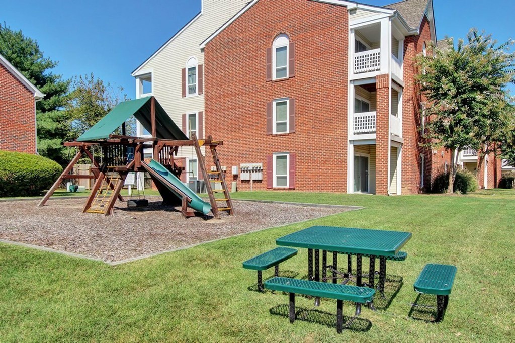 A playground with a green slide and picnic tables in front of a red brick apartment building.