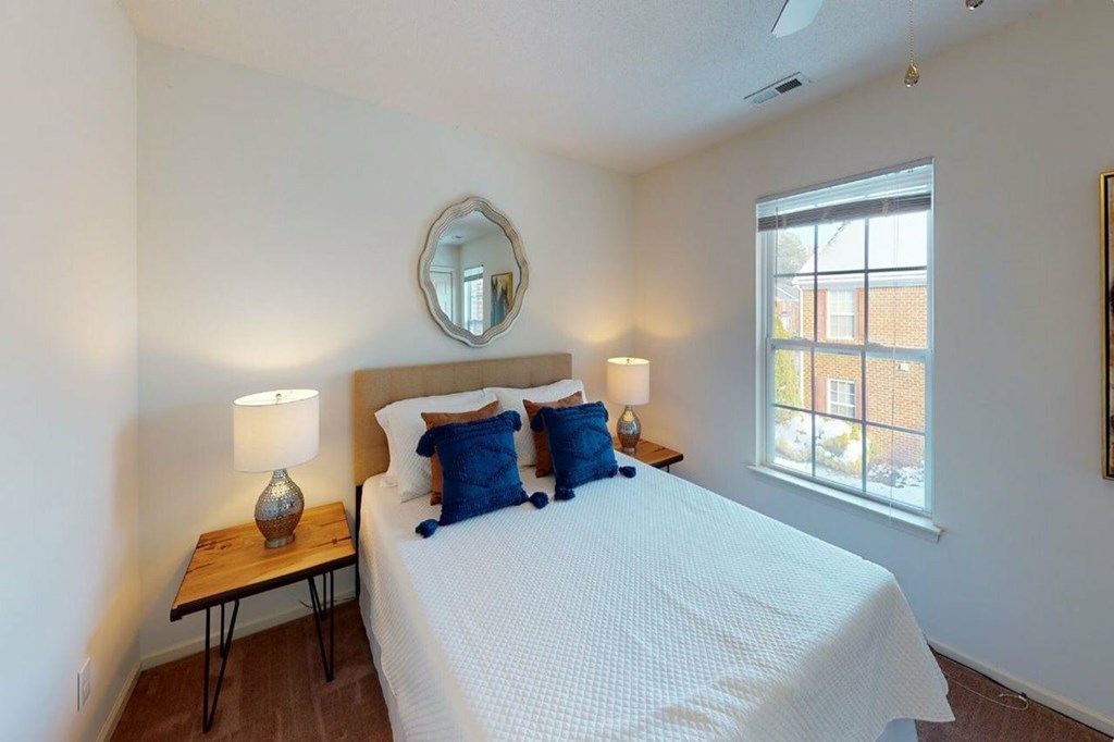 A bedroom with a bed, a table with a lamp, and a window.