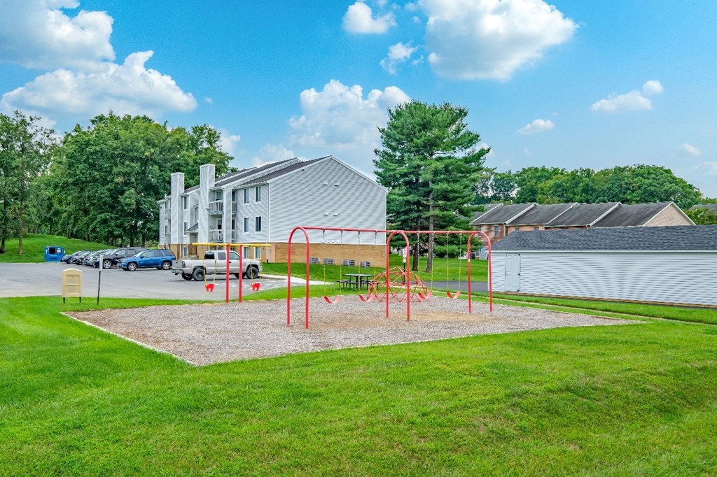A playground with a red swing set in front of a building and a parking lot.