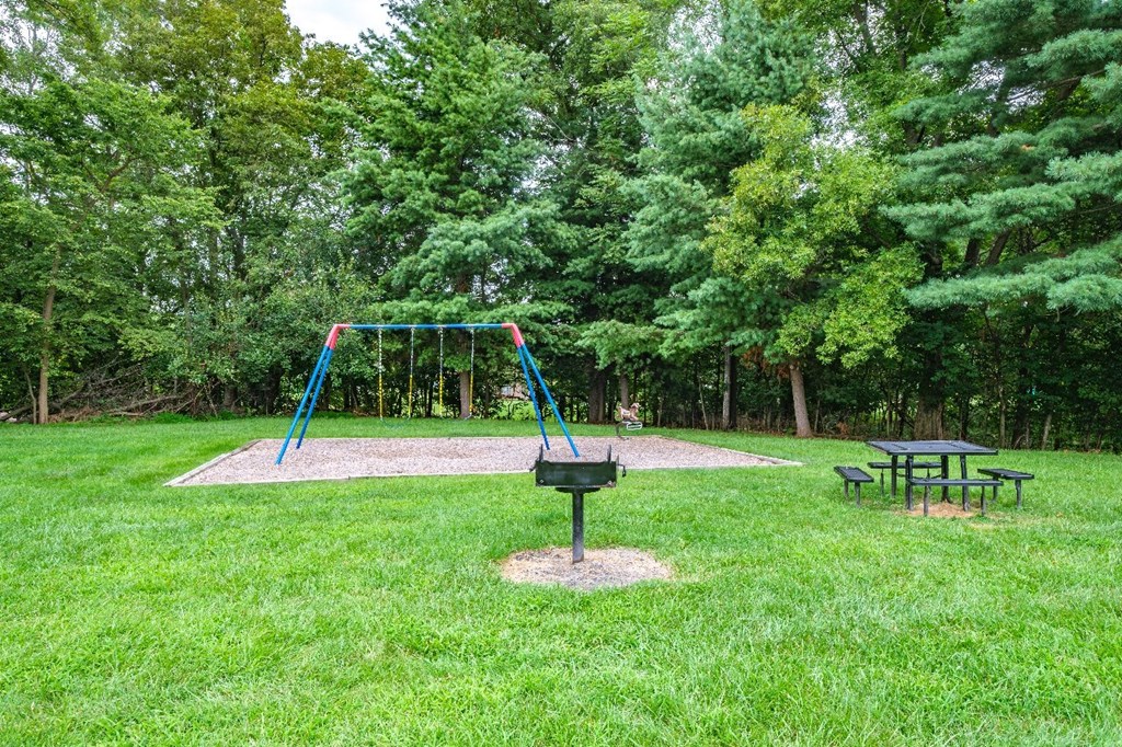 A playground with a swing set and picnic tables in a lush green park.