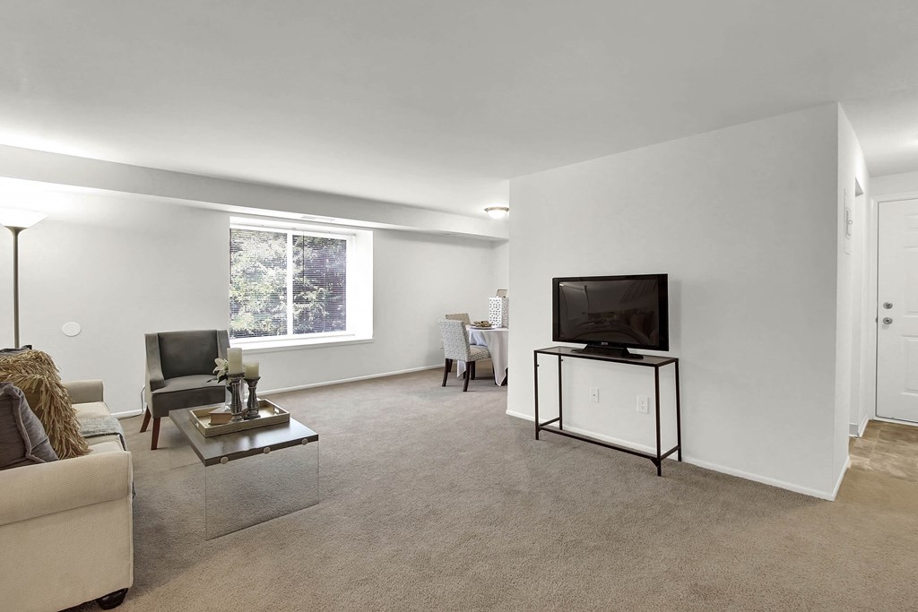 A living room with a white couch, a grey armchair, a glass coffee table, a flat screen TV on a stand, and a window with a view of trees.