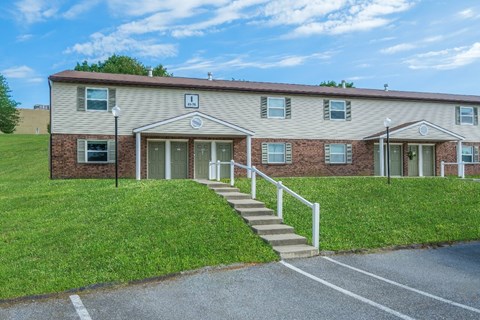 A building with a brown brick facade and a white door is surrounded by a grassy area and a paved driveway.