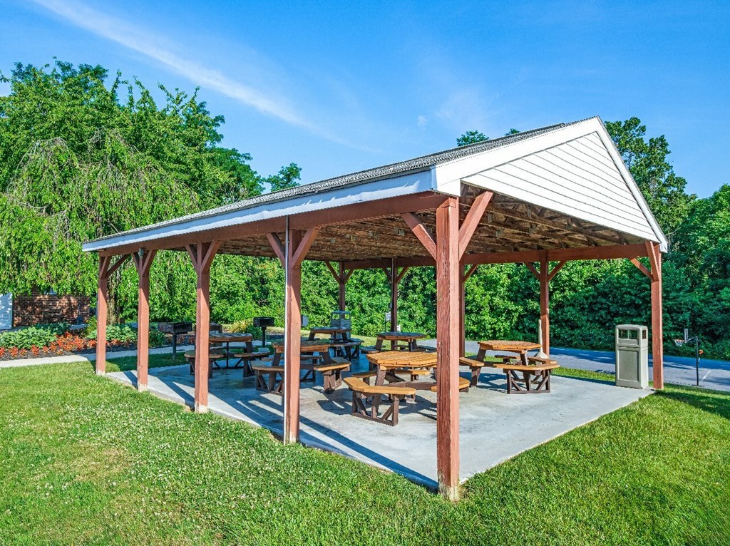A covered picnic area with tables and benches is surrounded by green grass and trees.