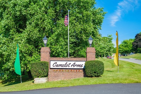 A sign that says "Camelot Arms" is in front of a green hedge.
