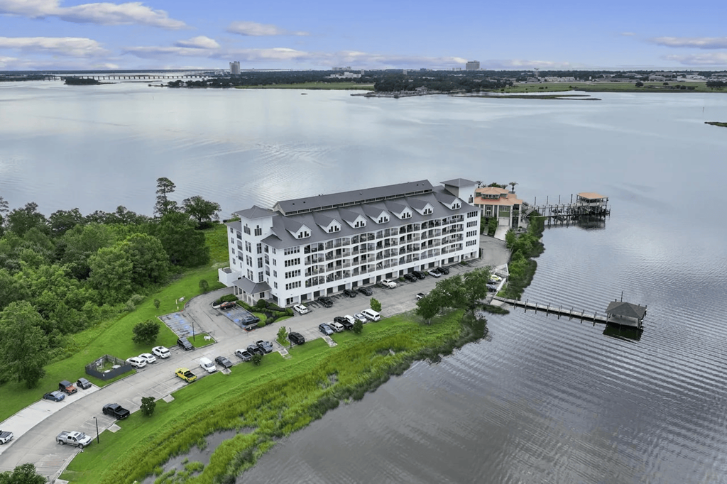 A large white building with a grey roof is surrounded by water and greenery.
