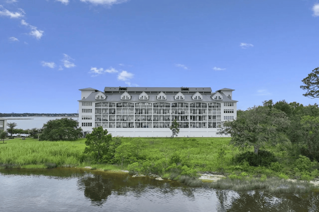 A large white building with a grey roof is surrounded by greenery and water.