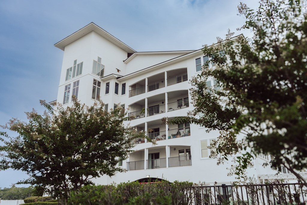 A white building with balconies and windows is surrounded by trees.