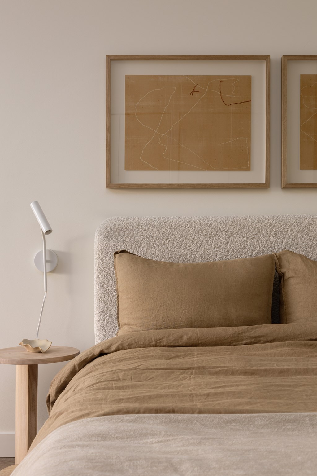 A bed with a tan comforter and two tan pillows sits in a room with two framed pieces of art on the wall.