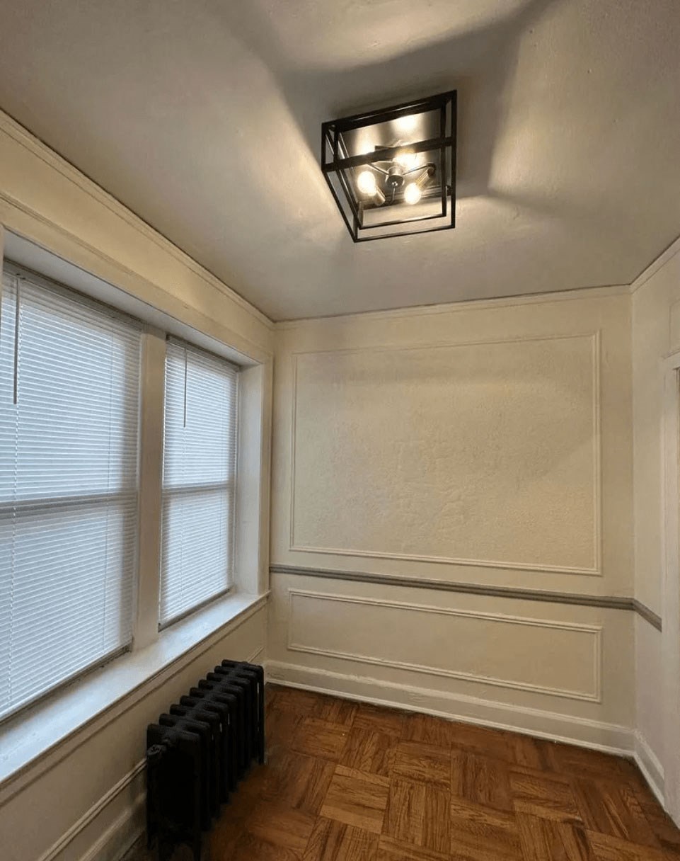 A room with a wooden floor, a window with blinds, a radiator, and a light fixture.