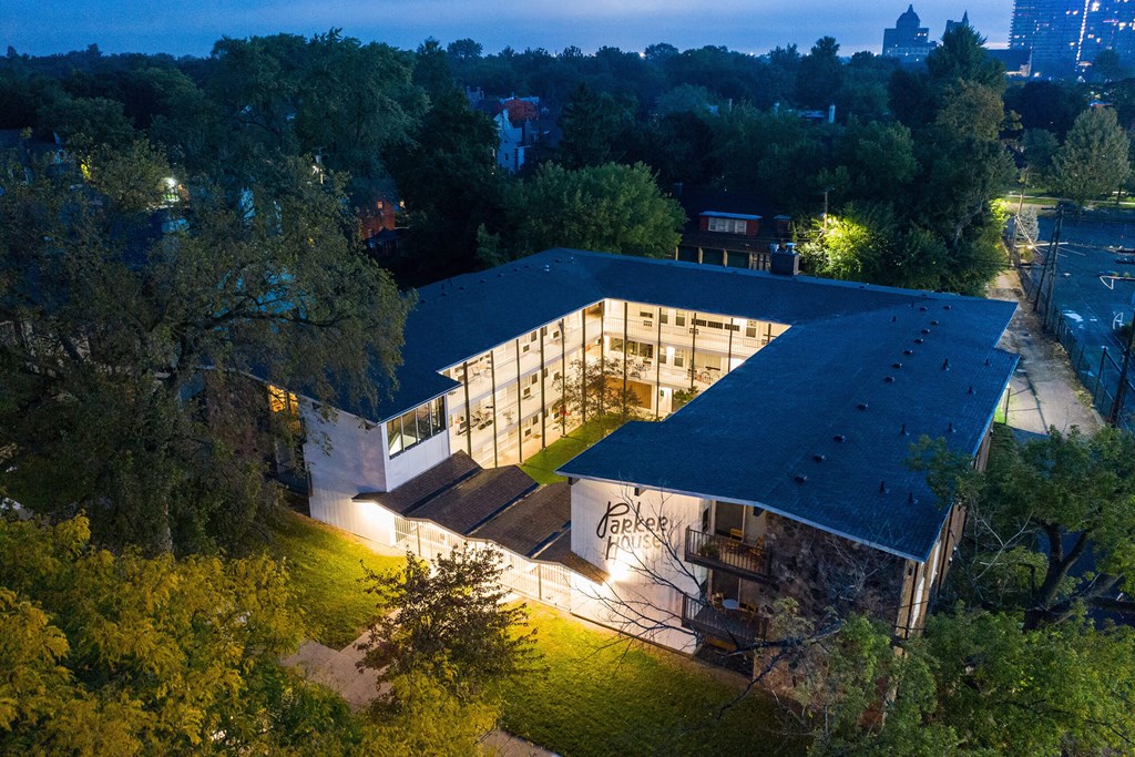 A modern building with a dark roof and lit windows is surrounded by trees.