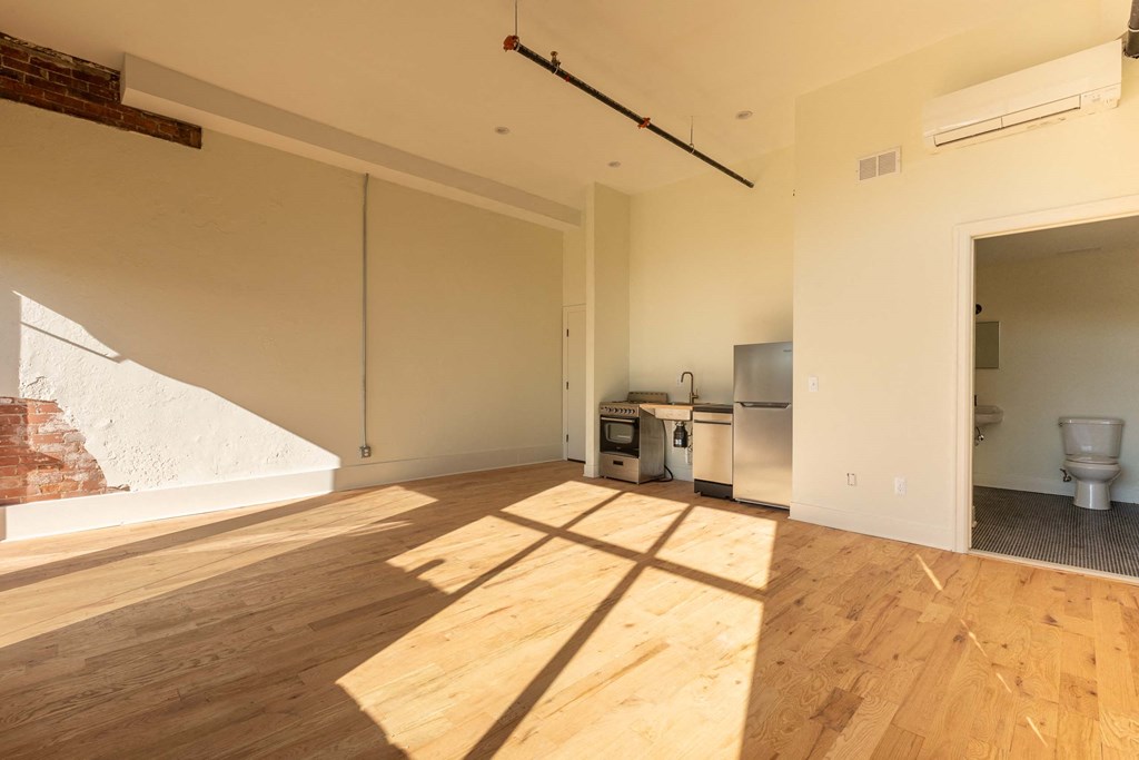 A room with wooden floors and white walls, with a kitchen area visible in the background.
