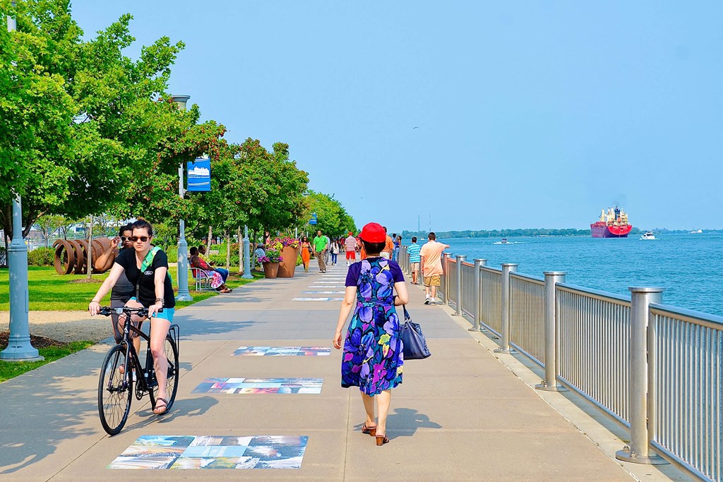 A woman in a floral dress walks down a sidewalk next to a body of water.