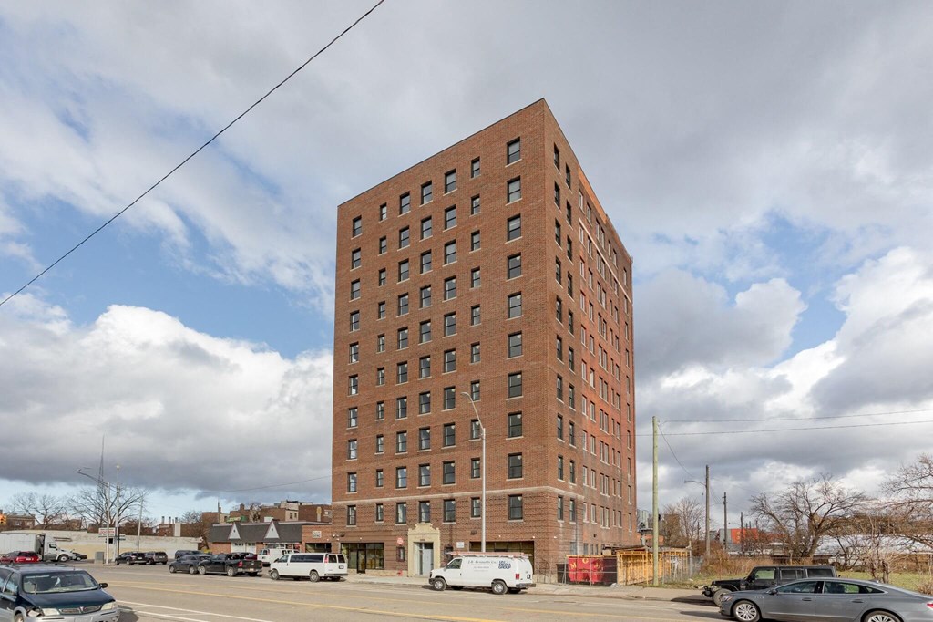 A tall red brick building with many windows is surrounded by cars and a cloudy sky.