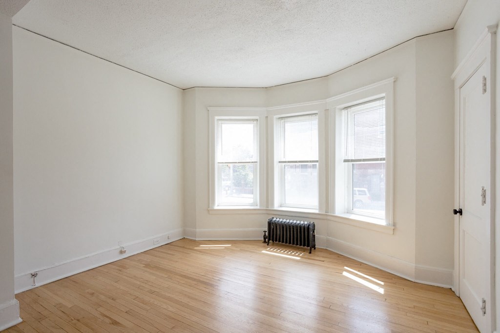 A room with wooden floors and white walls has a radiator and three windows.